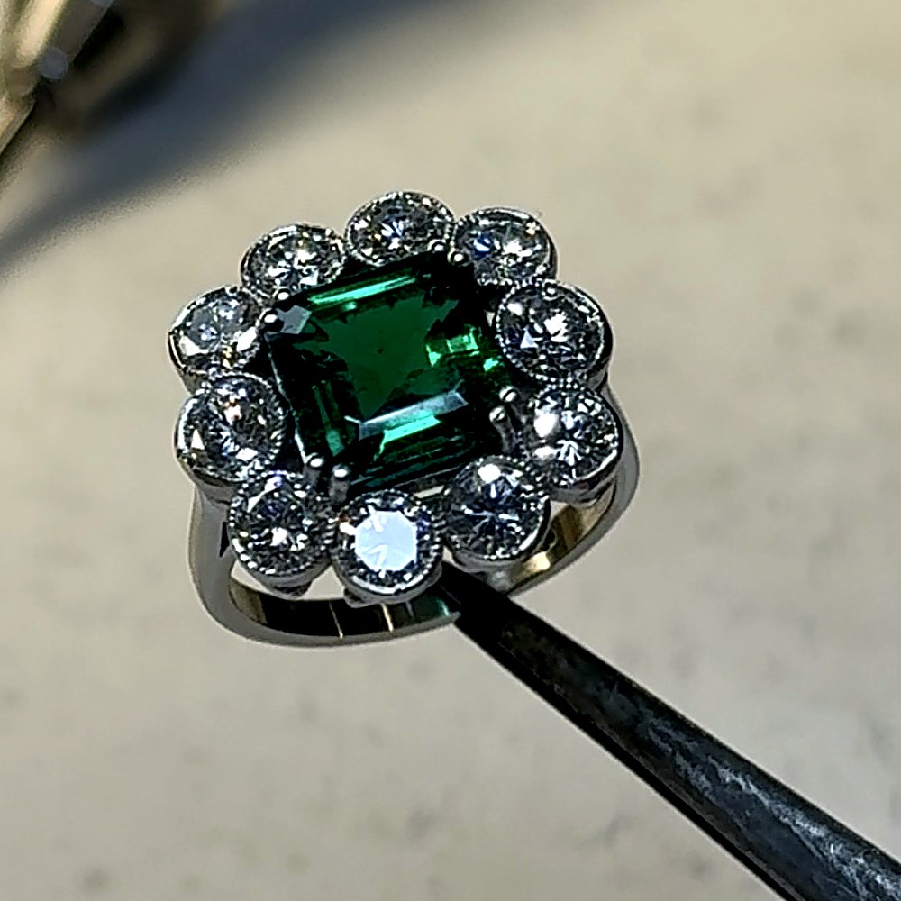 Emerald cut green emerald in a diamond cluster ring, grain set in platinum with a milgrain edge